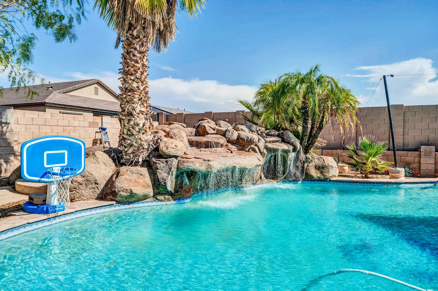 The backyard pool with a rock waterfall, palm trees and a basketball hoop.