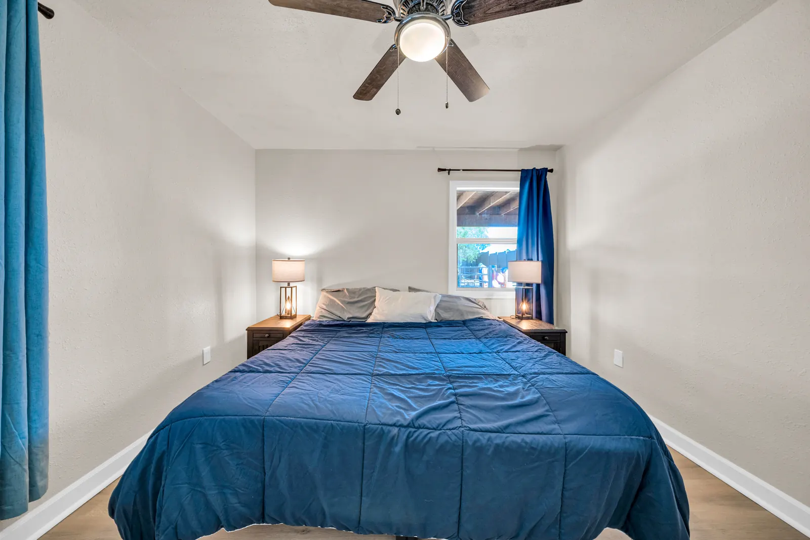 A bedroom with a made bed in a blue cover, lamps on both sides and a ceiling fan.