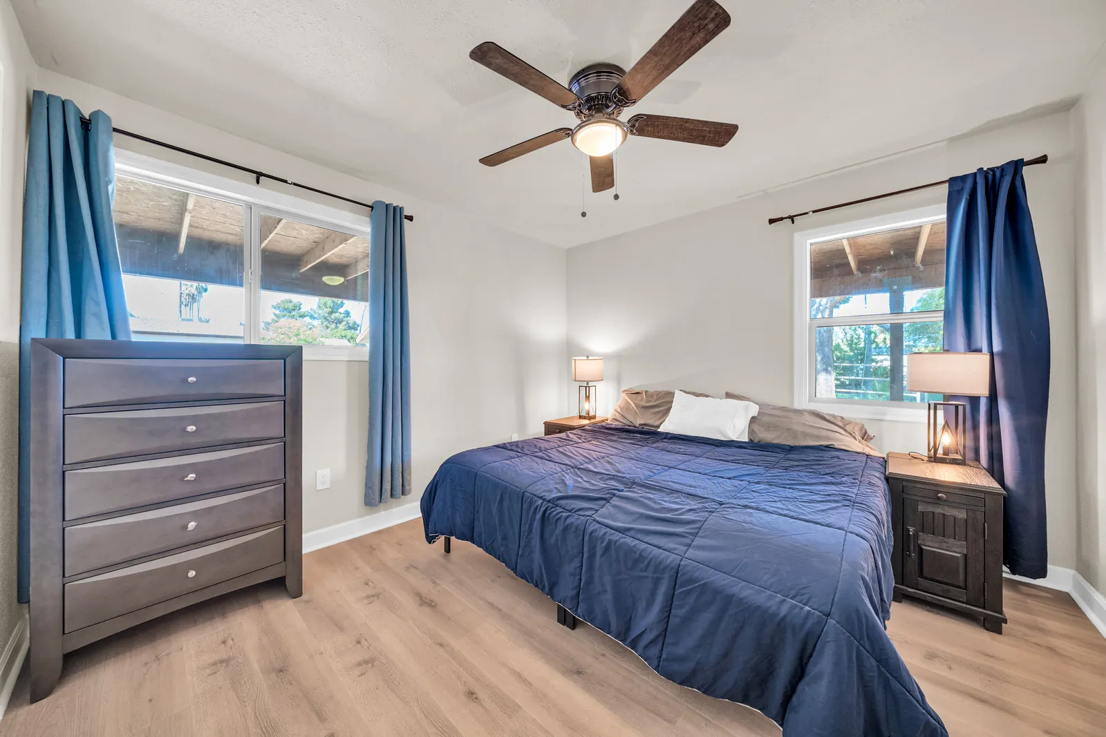 A second bedroom with a dresser, a bed with a navy cover and two windows.